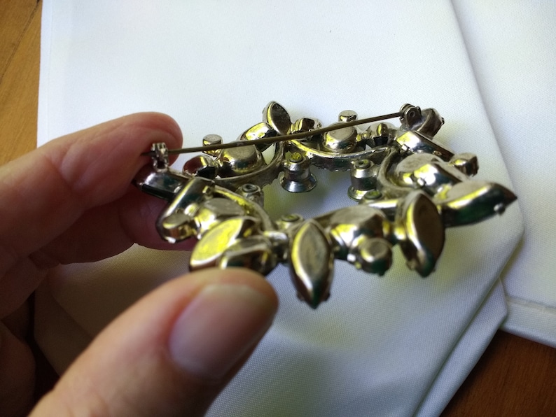 May include: Vintage silver-toned brooch with a floral design. The brooch features a wreath-like arrangement of clear and amber-colored rhinestones. The pin is held in place by a safety clasp.