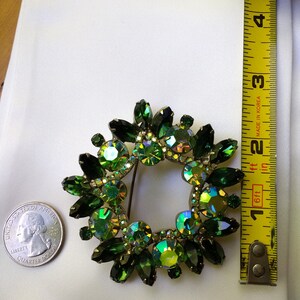 May include: A vintage green rhinestone brooch in a wreath design. The brooch features various shades of green stones, including emerald and iridescent hues. The brooch is approximately 2 inches in diameter. A US quarter is next to the brooch for size comparison.