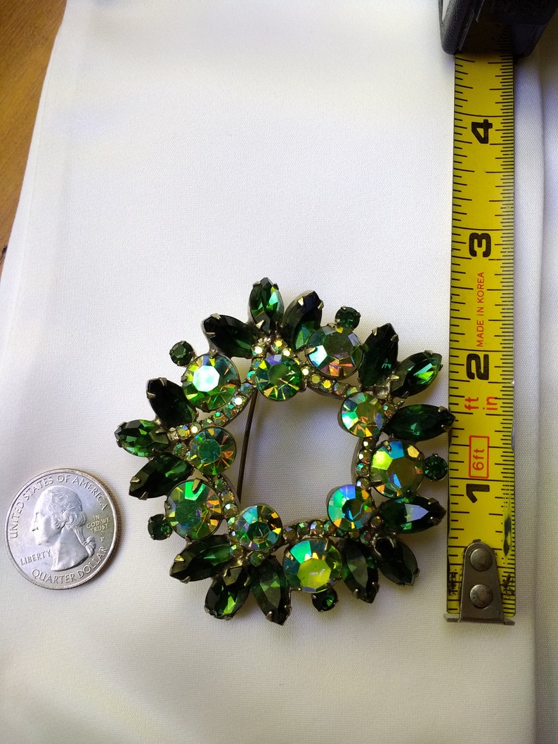 May include: A vintage green rhinestone brooch in a wreath design. The brooch features various shades of green stones, including emerald and iridescent hues. The brooch is approximately 2 inches in diameter. A US quarter is next to the brooch for size comparison.