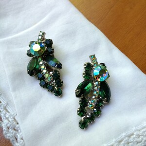 May include: A pair of vintage clip-on earrings featuring a cluster of green and iridescent rhinestones. The earrings have a silver-tone metal setting and are arranged in a leaf-like design. They rest on a white cloth.