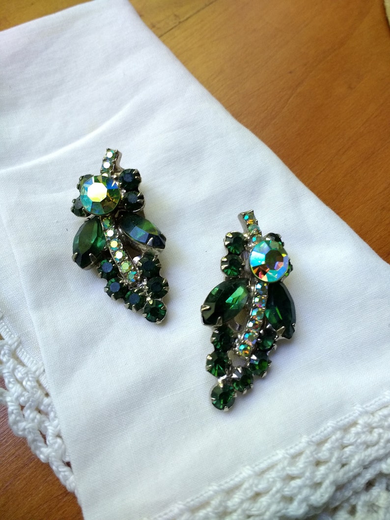 May include: A pair of vintage clip-on earrings featuring a cluster of green and iridescent rhinestones. The earrings have a silver-tone metal setting and are arranged in a leaf-like design. They rest on a white cloth.