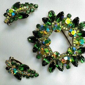 May include: A vintage jewelry set featuring a wreath-shaped brooch and matching clip-on earrings. The set is adorned with emerald green and iridescent rhinestones. The brooch has a pin closure. The earrings have a clip-on closure.