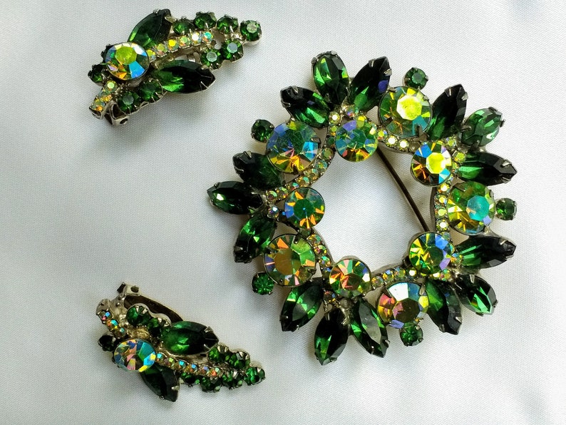 May include: A vintage jewelry set featuring a wreath-shaped brooch and matching clip-on earrings. The set is adorned with emerald green and iridescent rhinestones. The brooch has a pin closure. The earrings have a clip-on closure.