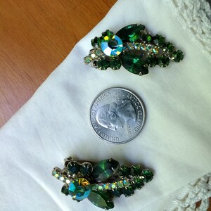 May include: Two vintage green rhinestone brooches. Each brooch features a cluster of green rhinestones and a larger, iridescent center stone. The brooches are displayed on a white cloth next to a quarter.