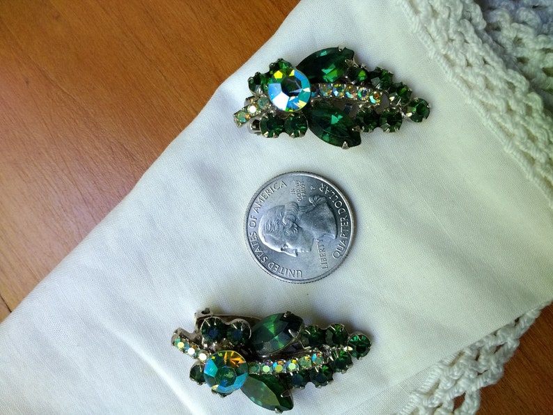 May include: Two vintage green rhinestone brooches. Each brooch features a cluster of green rhinestones and a larger, iridescent center stone. The brooches are displayed on a white cloth next to a quarter.