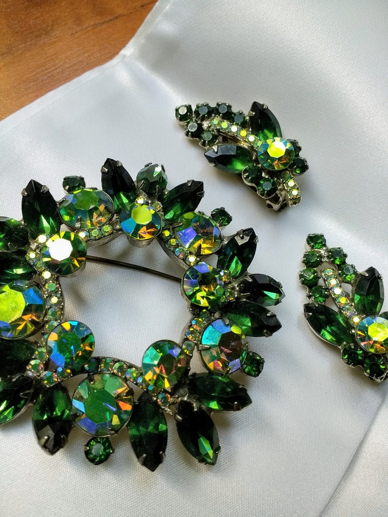 May include: A set of three green rhinestone brooches. The largest brooch is a circular wreath design. The other two brooches are smaller and have a leaf design.