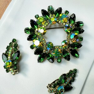 May include: A vintage jewelry set featuring a wreath-shaped brooch and matching clip-on earrings. The set is adorned with emerald green and iridescent crystals. The brooch is approximately 2 inches in diameter. The earrings are leaf-shaped.