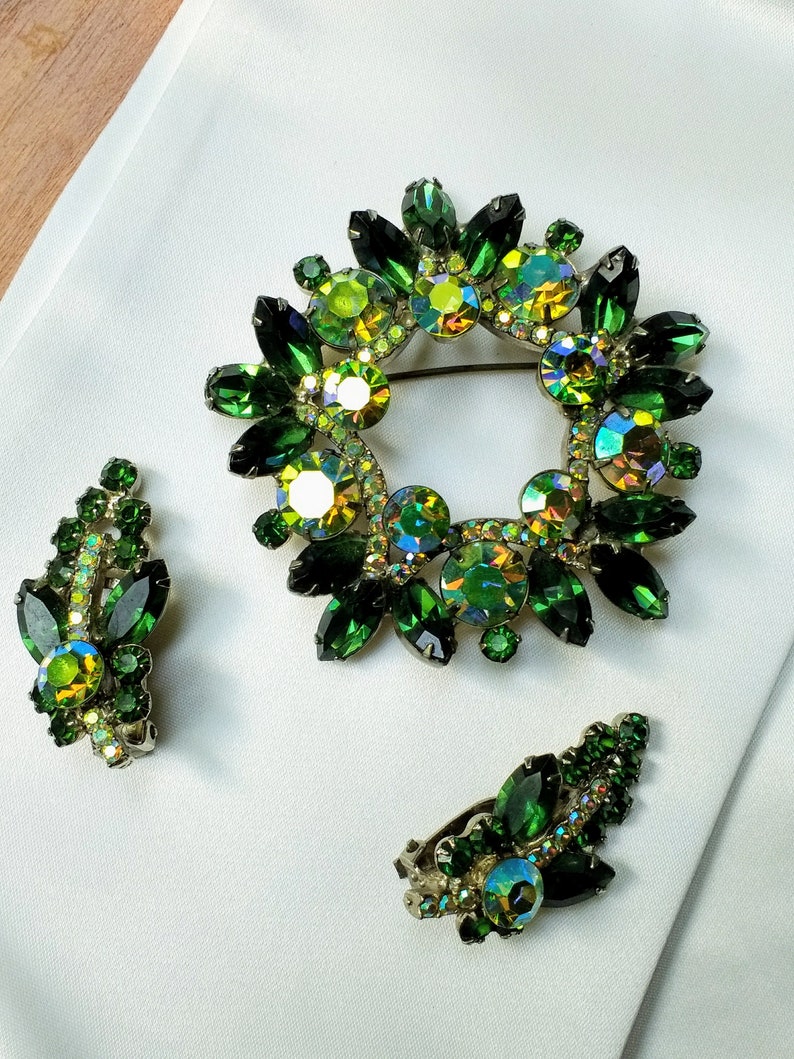 May include: A vintage jewelry set featuring a wreath-shaped brooch and matching clip-on earrings. The set is adorned with emerald green and iridescent crystals. The brooch is approximately 2 inches in diameter. The earrings are leaf-shaped.