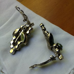 May include: A pair of vintage clip-on earrings. The earrings have a silver-tone metal setting with a cluster of clear and iridescent rhinestones. The earrings are resting on a white cloth.