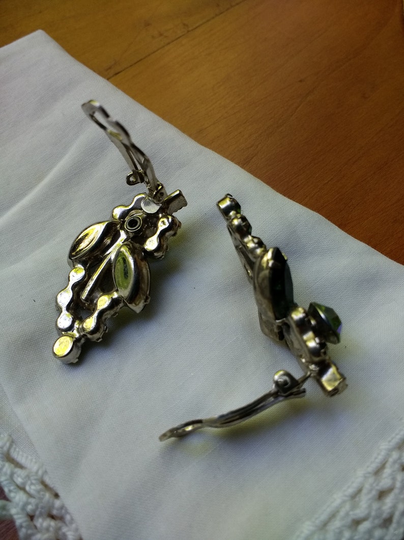 May include: A pair of vintage clip-on earrings. The earrings have a silver-tone metal setting with a cluster of clear and iridescent rhinestones. The earrings are resting on a white cloth.