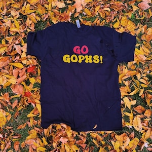 May include: A black t-shirt with the words "GO GOPHS!" in yellow and red letters on a bed of autumn leaves.