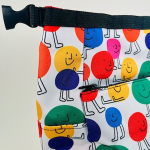 Puede incluir: Una bolsa de tela blanca con una cremallera negra y una correa negra. La bolsa est&aacute; cubierta con un patr&oacute;n de c&iacute;rculos sonrientes de colores con piernas.