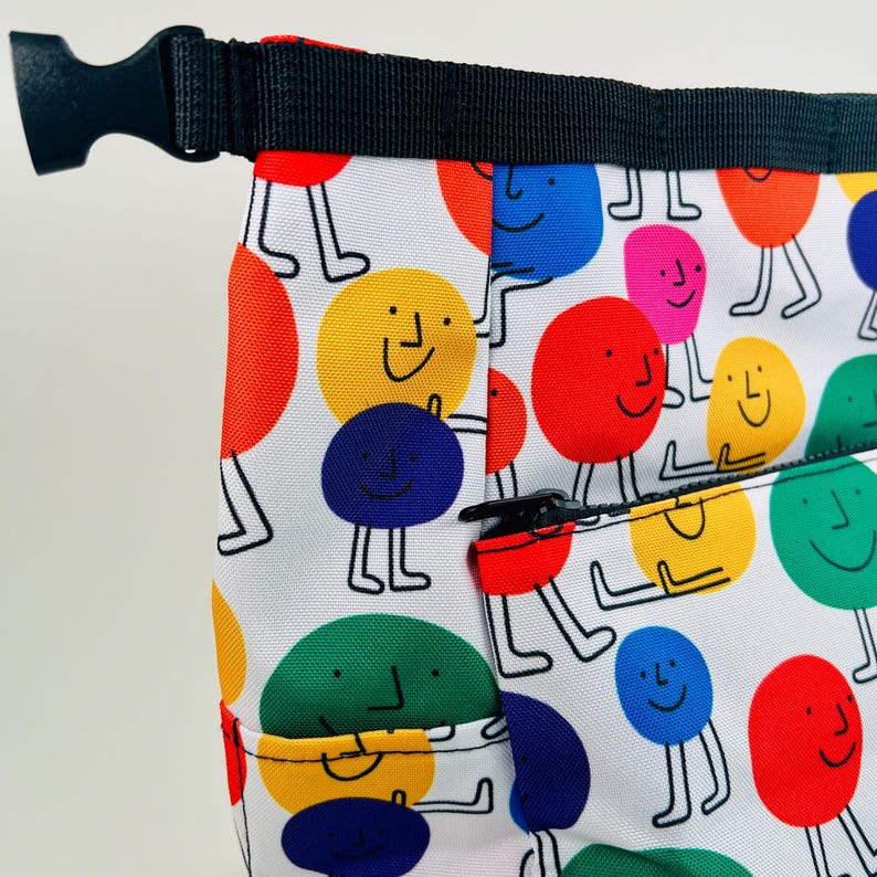 Puede incluir: Una bolsa de tela blanca con una cremallera negra y una correa negra. La bolsa est&aacute; cubierta con un patr&oacute;n de c&iacute;rculos sonrientes de colores con piernas.