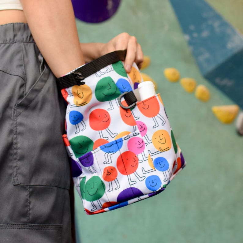 Puede incluir: Una bolsa de magnesio de escalada blanca con un patr&oacute;n colorido de caras sonrientes. La bolsa tiene una correa negra y un peque&ntilde;o bolsillo en el lateral.