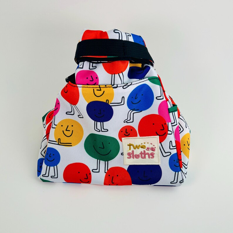 Puede incluir: Una bolsa de cord&oacute;n blanco con una correa negra y un patr&oacute;n colorido de caras sonrientes. La bolsa tiene una etiqueta rosa y blanca que dice "Twoca Sloths".