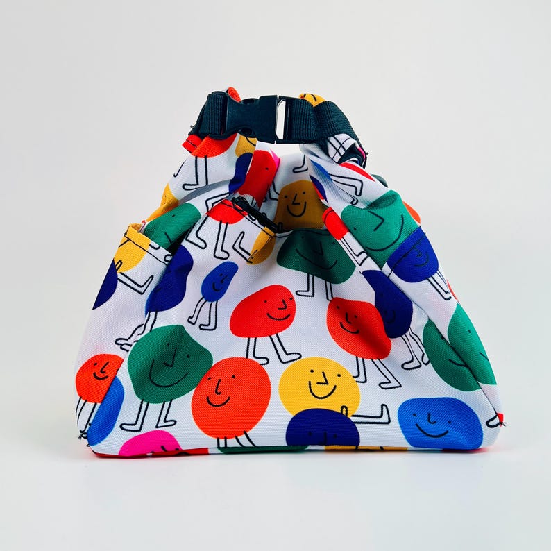 Puede incluir: Una bolsa de almuerzo blanca con un patr&oacute;n colorido de caras sonrientes. Las caras son de diferentes colores, incluyendo rojo, amarillo, azul, verde y rosa. La bolsa tiene una correa ajustable negra.