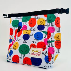 Puede incluir: Una bolsa de almuerzo blanca con una correa negra y un patr&oacute;n colorido de caras sonrientes. La bolsa tiene un bolsillo en la parte delantera con el texto "Two Sloths" en rosa.