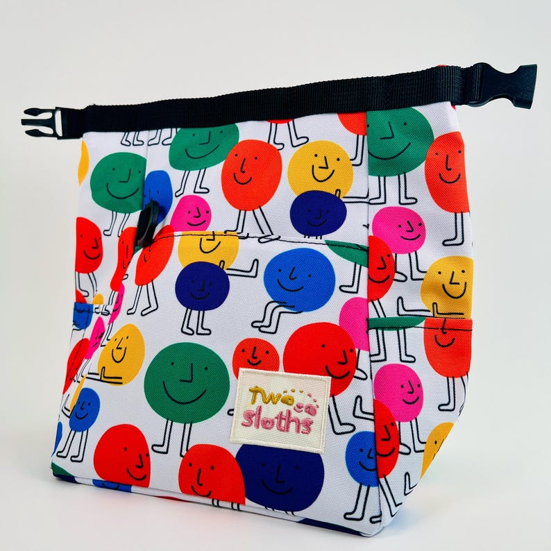 Puede incluir: Una bolsa de almuerzo blanca con una correa negra y un patr&oacute;n colorido de caras sonrientes. La bolsa tiene un bolsillo en la parte delantera con el texto "Two Sloths" en rosa.