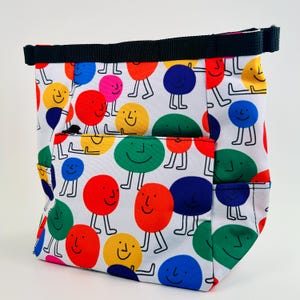 Puede incluir: Una bolsa de tela blanca con una correa negra y un patr&oacute;n colorido de caras sonrientes. Las caras son de diferentes colores y tienen diferentes expresiones.