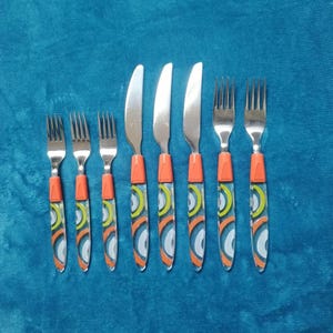May include: A collection of eight stainless steel cutlery items: four forks and four knives. The handles are orange with a clear overlay displaying a design of white, yellow, and blue concentric circles. The cutlery is arranged on a blue surface.