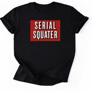 Op de afbeelding: Zwart t-shirt met de tekst "SERIAL SQUATER" in wit, omlijnd met zwart, op een rode rechthoekige achtergrond. Het shirt is opgevouwen, met opgerolde mouwen.