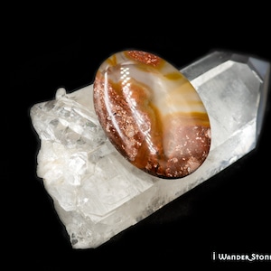 May include: A brown, white, and orange agate cabochon with a speckled pattern, resting on a clear quartz crystal.