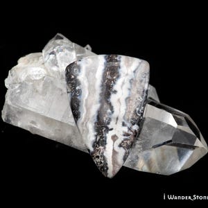 May include: A triangular cabochon stone featuring alternating bands of black, white, and translucent gray, resting against a clear, multifaceted quartz crystal cluster. The text "i Wander Stones and Jewelry" is printed in the bottom right corner.