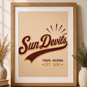 Può includere: Una stampa artistica incorniciata con il testo Sun Devils in un carattere corsivo marrone scuro con contorno dorato, su sfondo beige chiaro. Sotto il testo si legge TEMPE, ARIZONA e EST. 1885 in marrone, con un grafico a raggiera sopra.