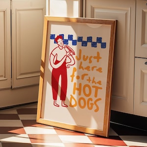 Op de afbeelding: Een ingelijste illustratie van een persoon die een hotdog eet met de tekst "Just here for the hot dogs" in oranje en gele letters.