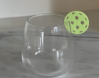 May include: A clear glass wine glass with a lime green pickleball on the rim.