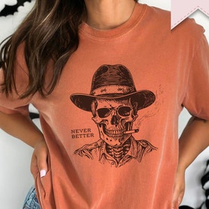 Puede incluir: Una camiseta naranja quemada con un gráfico de una calavera con un sombrero de vaquero y fumando un cigarrillo. El texto "Never Better" está impreso debajo de la calavera.