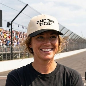 May include: A beige and black baseball cap with the text "START YOUR ENGINES" in black. The cap is worn by a person wearing a black t-shirt. The image is taken at a racetrack.