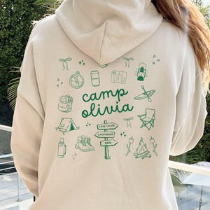 May include: A cream-colored hoodie with a green graphic design on the back. The design features a variety of camping-themed illustrations, including a tent, a compass, a lantern, a canoe, and a campfire. The text "camp olivia" is written in green letters.