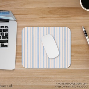 Puede incluir: Una alfombrilla de ratón rectangular con un patrón de rayas verticales en azul claro y crema. Un ratón de ordenador blanco descansa sobre la alfombrilla. Un portátil, un bolígrafo y una taza de café también son visibles en el escritorio de madera. El texto "* PATTERN PLACEMENT MAY VARY ON FINISHED PRODUCT" está en la parte inferior.