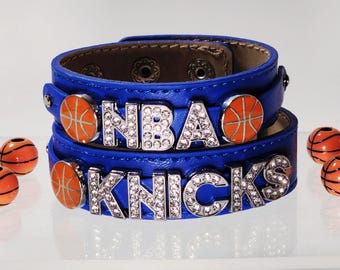 Może przedstawiać: Dwie niebieskie skórzane bransoletki typu wrap z metalowymi literami w kolorze srebrnym wysadzanymi przezroczystymi cyrkoniami, tworzącymi napis NBA i KNICKS. Każda bransoletka ma pomarańczowo-czarne zawieszki w kształcie piłek do koszykówki.