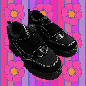 May include: A pair of black shoes with a chunky sole and a velcro strap closure. The shoes have white stitching details and a padded collar. The background features a colorful floral and striped pattern.