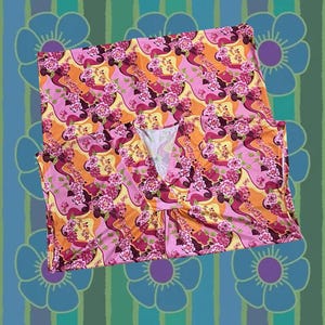 Könnte beinhalten: Ein ärmelloses Oberteil mit V-Ausschnitt, das ein lebhaftes Blumenmuster in Rosa, Orange und Gelb aufweist. Der Stoff ist mit einem sich wiederholenden Design aus stilisierten Blumen und organischen Formen verziert. Der Hintergrund zeigt ein ergänzendes Muster aus blauen und grünen Streifen und Blumenmotiven.