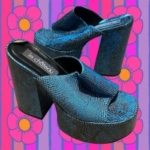 May include: A pair of blue platform sandals with a snakeskin pattern. The sandals have thick heels and a platform sole. The brand name "le château" is visible on the insole. The background features a colorful floral pattern.