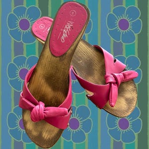 Peut inclure: Une paire de sandales roses avec des semelles en bois. Les sandales ont une conception de sangle nouée et la marque "Mossimo" est visible sur la semelle intérieure. Le fond présente un motif floral bleu et vert.