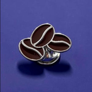 Peut inclure: Bague argentée ornée de trois breloques en forme de grains de café. Les grains sont brun foncé avec des détails argentés. La bague a une finition brillante, sur fond bleu. Accessoire sur le thème du café.