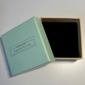 May include: A rectangular jewellery box with a light blue lid and a beige interior. The lid features the text "JEWELLERY - Especially For You" in black. The interior is lined with black fabric.