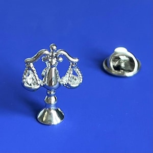 May include: A silver-toned metal pin featuring a detailed scale of justice design with clear rhinestones. The pin has a standard butterfly clasp on the back.