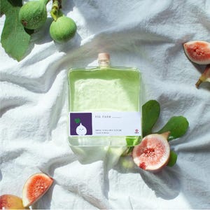 Puede incluir: Una botella cuadrada de vidrio transparente llena de líquido verde claro, rematada con un pequeño tapón de madera. La etiqueta blanca presenta el texto FIG FARM, BRING YOUR OWN NATURE, Natural Oil Diffuser y un cuadrado morado con un higo.
