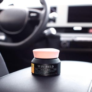 May include: A matte black cylindrical jar with a light pink lid. The label features a small orange graphic, the text YUZU FIELD, and the words citrus, sweet, fruity, earthy. The jar has a smooth finish and a wide base, presenting a clean design.