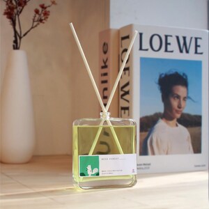 Op de afbeelding: Een vierkante glazen geurstokjesfles gevuld met lichtgele vloeistof, voorzien van twee witte rietjes en een wit etiket met een groene afbeelding van een eekhoorn en de tekst MOSS FOREST, BRING YOUR OWN NATURE, en Scent Oil Diffuser.