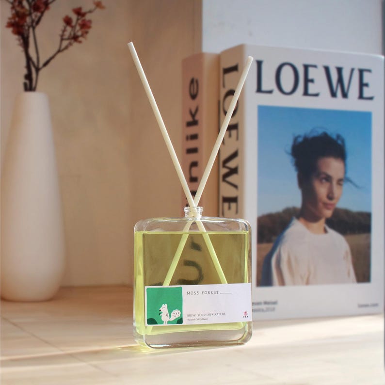 Op de afbeelding: Een vierkante glazen geurstokjesfles gevuld met lichtgele vloeistof, voorzien van twee witte rietjes en een wit etiket met een groene afbeelding van een eekhoorn en de tekst MOSS FOREST, BRING YOUR OWN NATURE, en Scent Oil Diffuser.