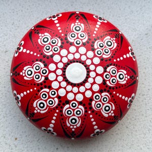 Könnte beinhalten: Ein runder, rot bemalter Stein mit einem Mandala-Design. Das Mandala weist weiße und schwarze Punktmuster und blütenartige Formen auf, die von einem zentralen weißen Kreis ausgehen. Der Stein ist ein dekorativer Gegenstand.