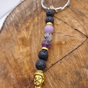 Puede incluir: Un llavero con un dije de Buda dorado, cuentas de roca volcánica negra y cuentas de piedras preciosas moradas, rosadas y transparentes.