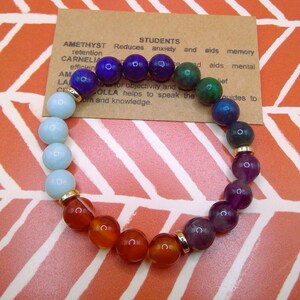 Puede incluir: Una pulsera de cuentas con una variedad de piedras preciosas de colores, incluyendo piedras azules, moradas, naranjas y verdes. La pulsera tiene espaciadores dorados y una pequeña tarjeta con texto sobre las propiedades de las piedras. El fondo es un diseño naranja y blanco.