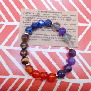Peut inclure: Un bracelet chakra arc-en-ciel avec des perles dorées séparant chaque couleur. Le bracelet est composé de perles d'œil de tigre, de cornaline, de jaspe rouge, d'améthyste, de sodalite, de lapis-lazuli et de fluorite. Le bracelet est sur un fond à chevrons rose et blanc avec une carte qui indique "FLUORITE SCIENTIST, ENGINEERS the genius stone, helps boosts advancement mentally, physically and spiritually AMETHYST helps develop ability to think clearly and not clouded w CARNELIAN efficie TIGERS concentra to stay focused, to d aids mental motivation, unity and focus uition sis SOE Student's Stone' helps with i manizational abilities, good for enha personal perception, enhances"